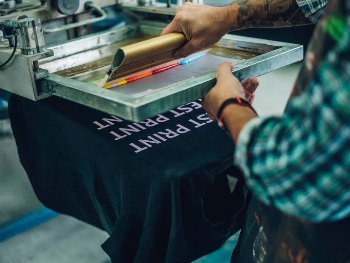 Siebdruck auf ein dunkles T-Shirt: Eine Rakel zieht Farbe über das Sieb
