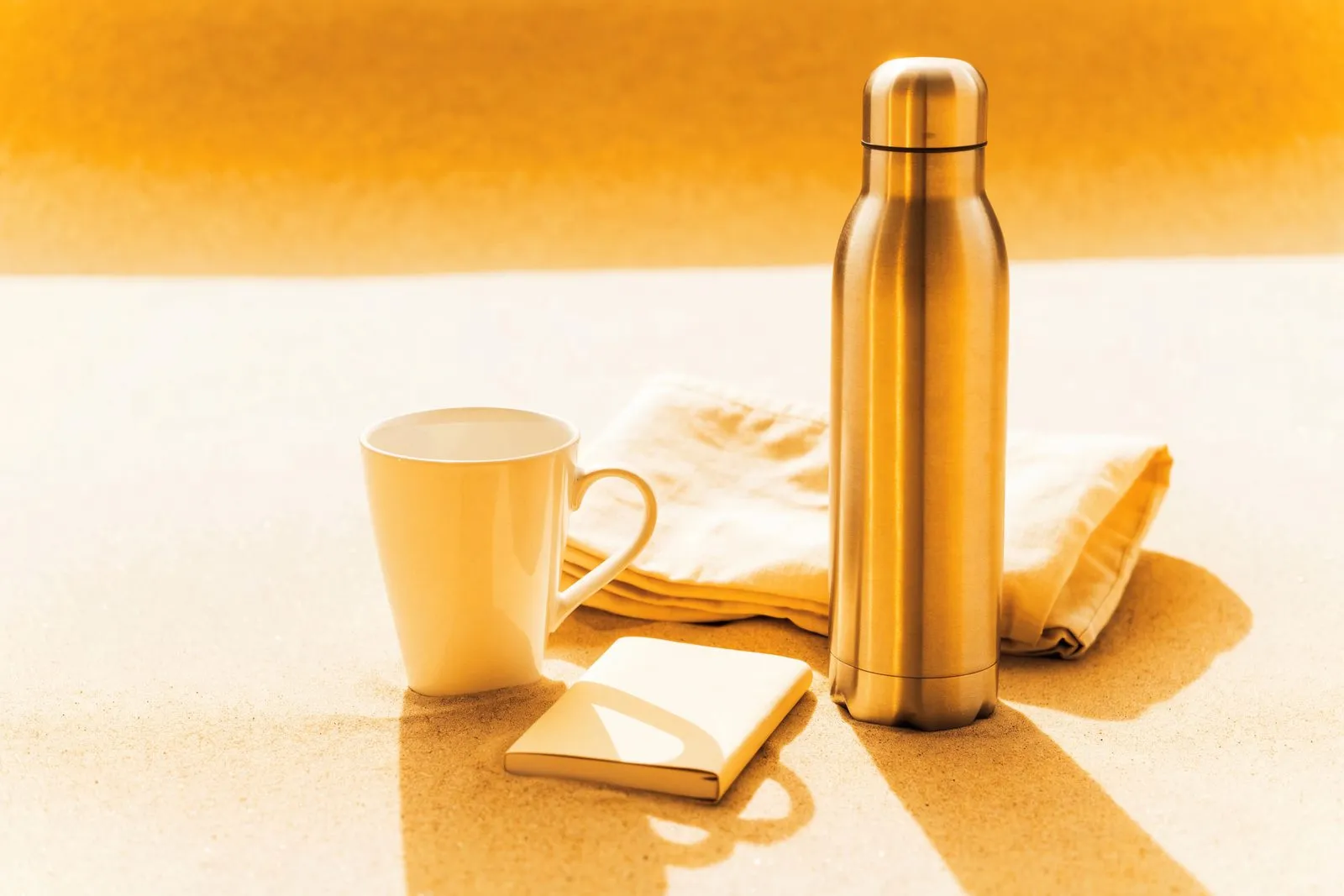 Unbedruckte Werbeartikel auf sandfarbenem Grund: Tasse, Trinkflasche, Baumwolltasche und Notizbuch