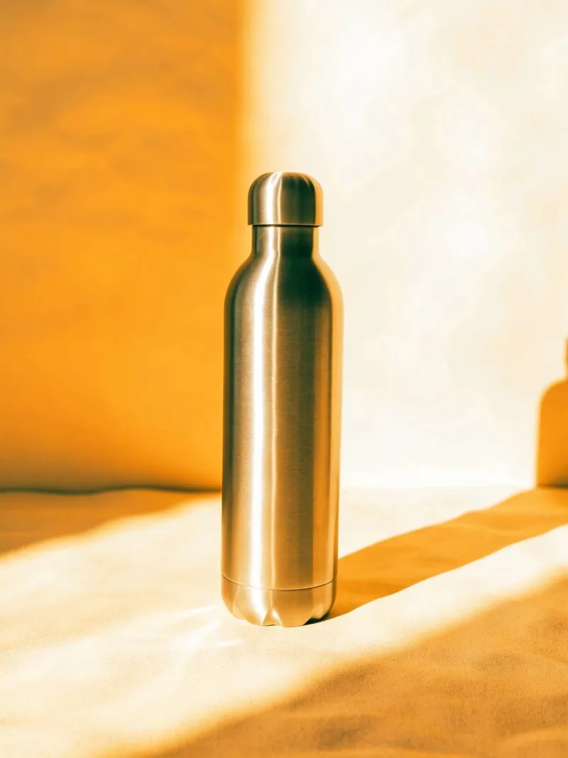 Schlichte Edelstahl-Trinkflasche steht im warmen Nachmittagslicht auf sandfarbenem Grund